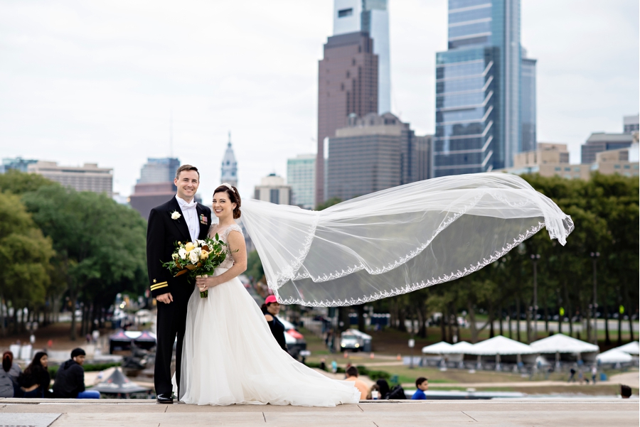 A wedding couple pose at the top of the Philadelphia Art Museum while the brides veil blows in the wind.